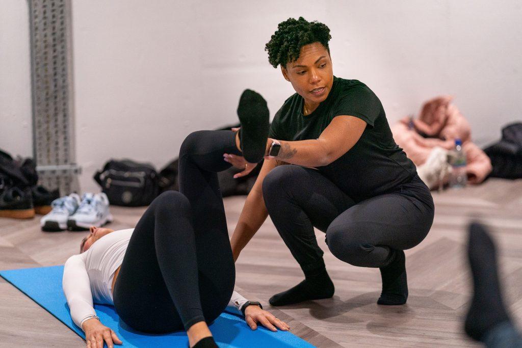 Are Pilates good for weight loss? One of our coaches supporting a client as she lays on a yoga mat with one leg bent and another raised at 90 degrees. The coach holds this in place for her.
